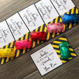 May include: Five small toy cars in different colors, including yellow, pink, red, green, and blue, are placed on top of cards with the text "I'm wheelic GLAD WE ARE friends! Love, Evie" printed on them. The cards have a black and yellow striped background.