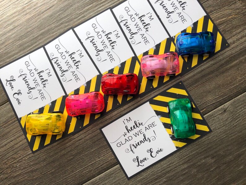 May include: Five small toy cars in different colors, including yellow, pink, red, green, and blue, are placed on top of cards with the text "I'm wheelic GLAD WE ARE friends! Love, Evie" printed on them. The cards have a black and yellow striped background.