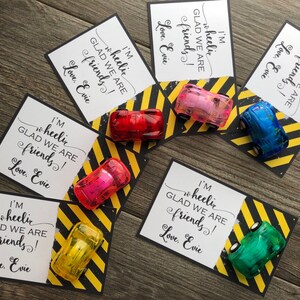 May include: Five small toy cars in different colors are placed on top of cards with a black and yellow striped background. The cards have the text "I'm wheelic glad we are friends! Love, Evie" printed on them.