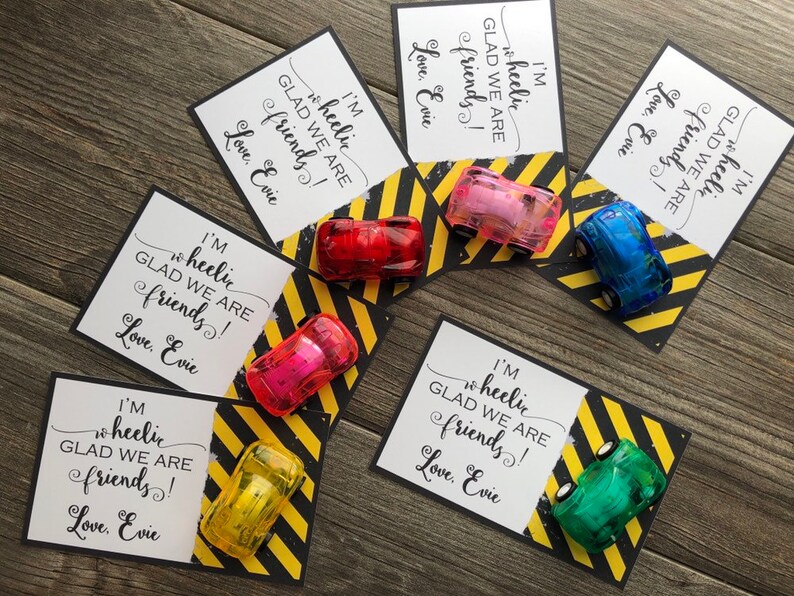 May include: Five small toy cars in different colors are placed on top of cards with a black and yellow striped background. The cards have the text "I'm wheelic glad we are friends! Love, Evie" printed on them.