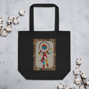May include: A black tote bag with a colorful illustration of a Native American Kachina doll. The doll is wearing a red and blue outfit and has a yellow sun symbol on its head. The background is a beige color with a brown border.