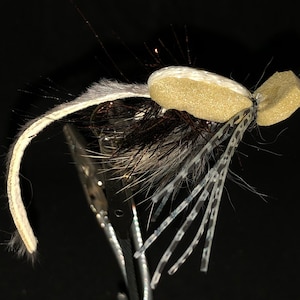 May include: A close-up of a handmade fishing fly. The fly features a tan foam body, black and gray feathers, and white and gray patterned legs. The fly is held by a silver clamp against a black background.