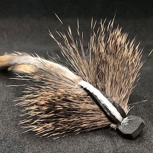 May include: A handmade fly fishing lure with a dark gray body and a cluster of brown and gray feathers. The lure has a white stripe and a small, dark gray head. The background is black.