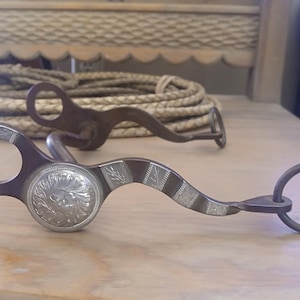 May include: Two antique horse bits with decorative silver accents. One bit features a floral design on the center plate and striped patterns. The other bit is a simpler design. A braided rope is in the background.