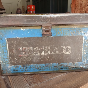 May include: A rectangular metal bread box featuring a distressed blue and brown finish with visible paint chipping. The front panel displays the word BREAD in large, weathered white capital letters centered within a rectangular brown border.