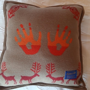 May include: A square tan throw pillow featuring two orange and red handprint graphics in the center, red deer silhouettes along the bottom, and red lizard motifs in the corners. A blue Pendleton brand label is stitched on the bottom right corner.