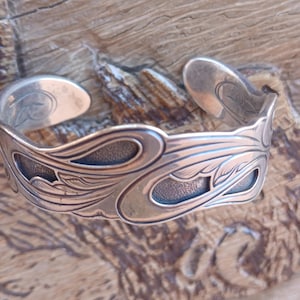 May include: A silver cuff bracelet featuring an intricate, raised floral and leaf pattern with recessed, darkened areas that provide contrast. The metal has a polished finish, and the design wraps around the band, which has open ends.