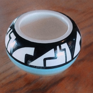 May include: A small, round ceramic bowl featuring a smooth, white interior and a glossy exterior. The exterior is decorated with a bold, black and white geometric pattern consisting of sharp, angular lines and shapes encircling the entire vessel.