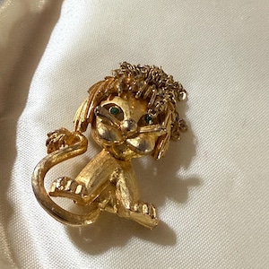 May include: A gold-toned lion brooch with green gemstone eyes. The lion is depicted in a seated position with a textured mane and tail. The brooch is set against a white background.