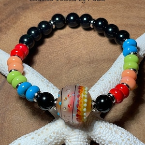 Puede incluir: Una pulsera de cuentas coloridas con una cuenta central estampada. La pulsera incluye cuentas negras, rojas, naranjas, verdes y azules, separadas por espaciadores plateados. El texto "Seaside Jewels by Paula" es visible. La pulsera está sobre una estrella de mar blanca.