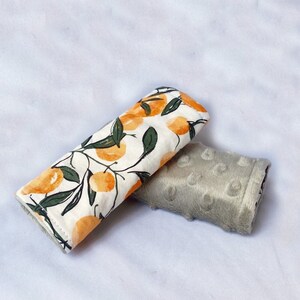May include: Two fabric stroller straps, one with a white background and a pattern of orange citrus fruit and green leaves, and the other a solid light gray with a textured surface.