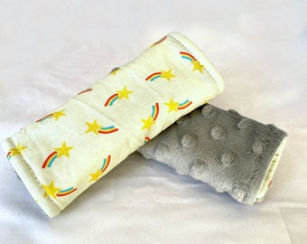 May include: Two car seat strap covers. One is white with yellow stars and rainbow designs. The other is gray with a textured surface.