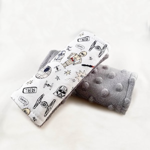 May include: A white fabric car seat strap cover with a Star Wars theme. The cover features a black and white illustration of Darth Vader, R2-D2, C-3PO, and X-Wing fighters. The text "Rebel" appears on the cover. The cover is paired with a gray, textured, plush car seat strap cover.
