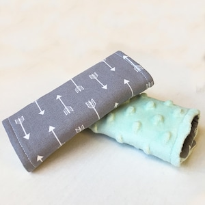 May include: Two gray and mint green car seat belt covers. One cover has a white arrow pattern and the other has a textured surface.