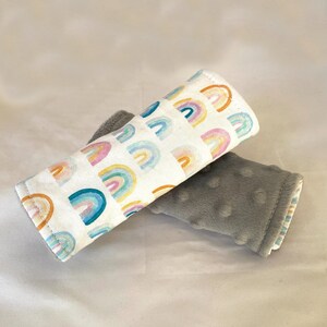 May include: Two fabric baby stroller straps. One strap is white with a rainbow pattern and the other is gray with a textured surface.