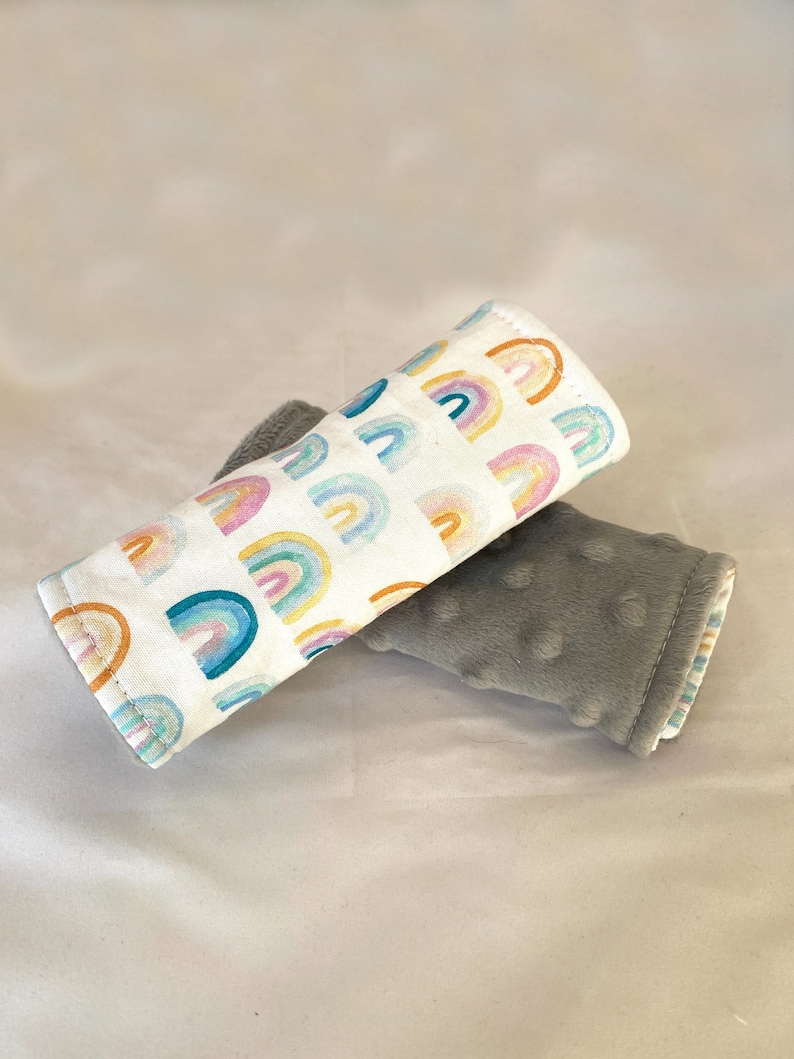 May include: Two fabric baby stroller straps. One strap is white with a rainbow pattern and the other is gray with a textured surface.