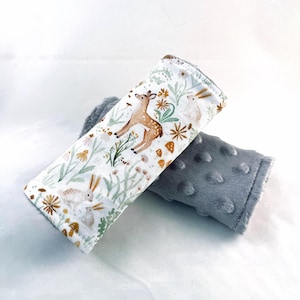 May include: Two baby seatbelt covers. One is white with a woodland print of a deer, rabbits, and flowers. The other is gray, soft, and textured. The covers are designed to protect a child's neck from seatbelt chafing.