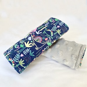 May include: A blue and green fabric car seat strap cover with a dinosaur and rainbow pattern. The cover is made of soft, plush fabric and is designed to protect car seats from wear and tear.