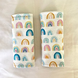 May include: Two white fabric car seat belt covers with a rainbow pattern. The covers are made of a soft, comfortable fabric and are designed to protect your car seats from wear and tear.