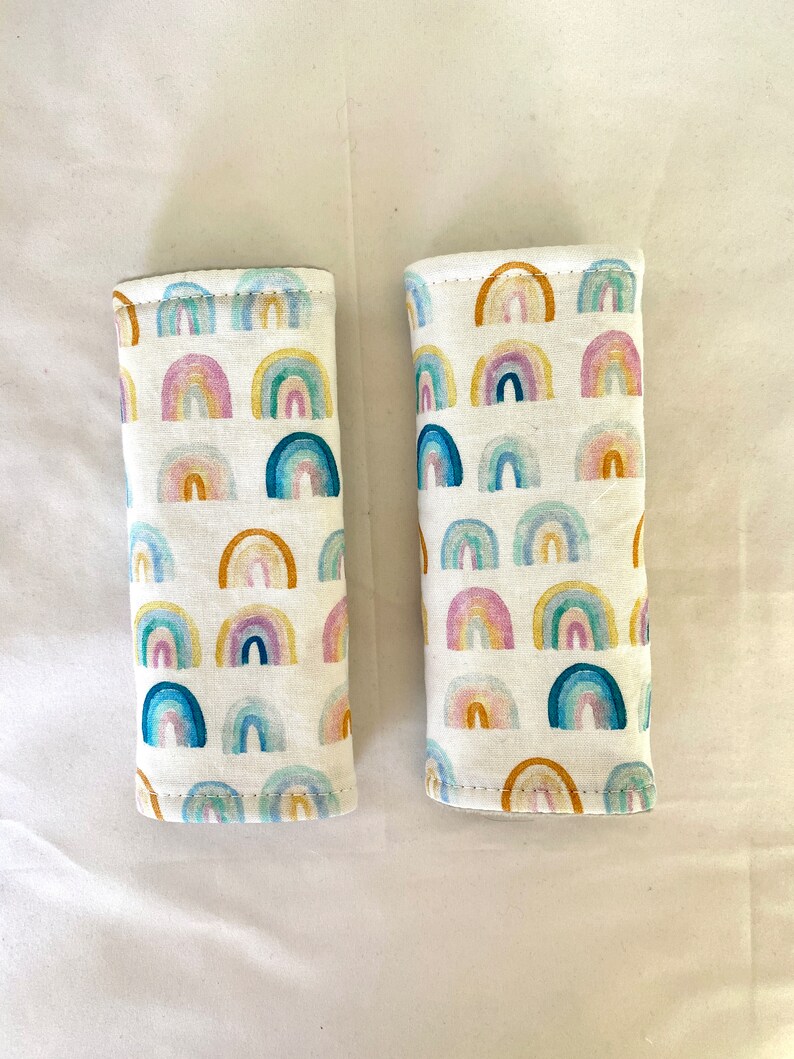 May include: Two white fabric car seat belt covers with a rainbow pattern. The covers are made of a soft, comfortable fabric and are designed to protect your car seats from wear and tear.