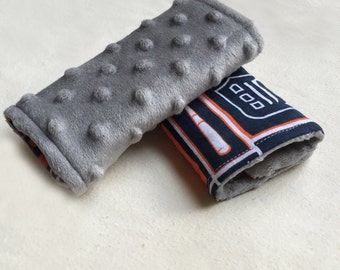 May include: Two gray and white fabric stroller straps. One strap has a gray, textured surface and the other has a navy blue, orange, and white pattern with a baseball bat and a home plate graphic.