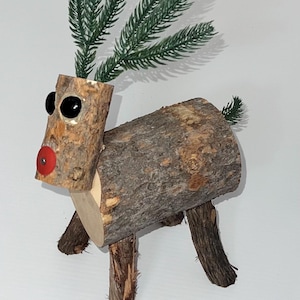 May include: A handcrafted wooden reindeer figurine with a rustic aesthetic. The reindeer features a log body, twig legs, and a head made from a section of a log. It has black eyes, a red nose, and green faux pine needles for antlers and tail.