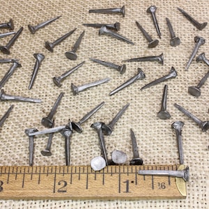 May include: A collection of 40 antique iron nails with flat heads, scattered on a burlap surface. A wooden ruler is placed below the nails, showing measurements in inches.