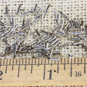 Puede incluir: Primer plano de un mont&oacute;n de tachuelas o clavos de tapicer&iacute;a de color plateado sobre un fondo de arpillera. Una regla se coloca debajo de las tachuelas, mostrando medidas en pulgadas.