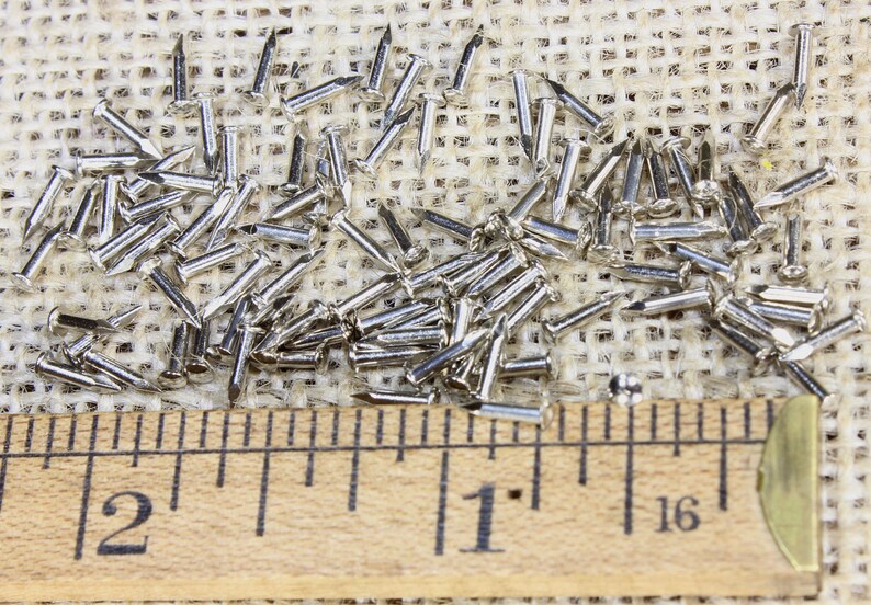 Puede incluir: Primer plano de un mont&oacute;n de tachuelas o clavos de tapicer&iacute;a de color plateado sobre un fondo de arpillera. Una regla se coloca debajo de las tachuelas, mostrando medidas en pulgadas.