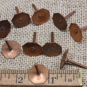 May include: Ten copper tacks with octagonal heads and a nail-like shank. The tacks are arranged in a random pattern on a brown hessian surface. A ruler is visible in the bottom of the image.