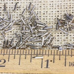 Puede incluir: Primer plano de un mont&oacute;n de tachuelas plateadas o clavos de tapicer&iacute;a sobre una superficie de arpillera. Una regla de madera con marcas en pulgadas se coloca debajo de las tachuelas. La regla muestra las medidas de 1 pulgada y 2 pulgadas.