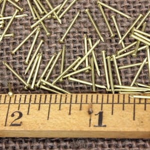 May include: A close-up of a pile of small, gold-colored tacks or brads scattered on a brown burlap surface. A wooden ruler with inch markings is placed below the tacks, showing the tacks are about 1 inch long.