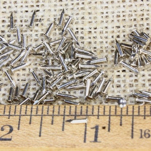 Puede incluir: Primer plano de un mont&oacute;n de tachuelas plateadas o clavos de tapicer&iacute;a sobre una superficie de arpillera. Una regla de madera con marcas en pulgadas se coloca debajo de las tachuelas. La regla muestra las medidas 1 y 2 pulgadas.