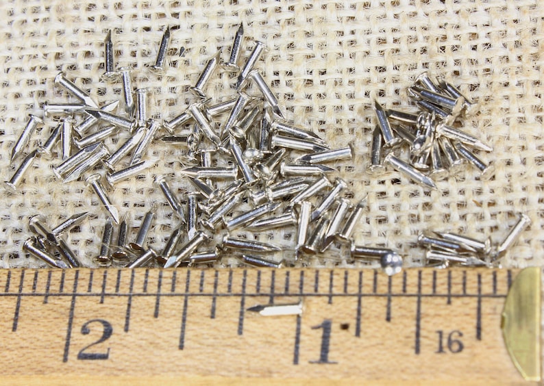 Puede incluir: Primer plano de un mont&oacute;n de tachuelas plateadas o clavos de tapicer&iacute;a sobre una superficie de arpillera. Una regla de madera con marcas en pulgadas se coloca debajo de las tachuelas. La regla muestra las medidas 1 y 2 pulgadas.