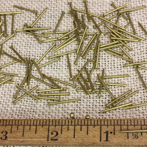 K&ouml;nnte beinhalten: Nahaufnahme eines Haufens von 2,5 cm langen Messingtackern auf einer Jutestoffoberfl&auml;che. Ein Lineal ist unter den Tackern platziert, um die Gr&ouml;&szlig;e zu zeigen.