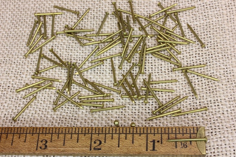 K&ouml;nnte beinhalten: Nahaufnahme eines Haufens von 2,5 cm langen Messingtackern auf einer Jutestoffoberfl&auml;che. Ein Lineal ist unter den Tackern platziert, um die Gr&ouml;&szlig;e zu zeigen.