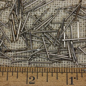 May include: A close-up of a pile of silver-colored tacks or brads on a burlap surface. A wooden ruler with inch markings is placed below the tacks.