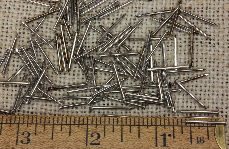 May include: A close-up of a pile of silver-colored tacks or brads on a burlap surface. A wooden ruler with inch markings is placed below the tacks.