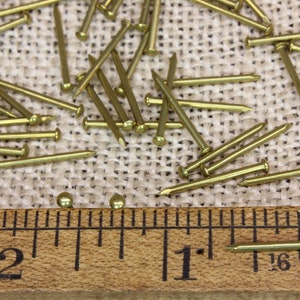 K&ouml;nnte beinhalten: Nahaufnahme eines Lineals, das 5 cm misst, mit einer Reihe kleiner, goldfarbener Rei&szlig;n&auml;gel, die auf einer Jutestoffoberfl&auml;che verstreut sind.