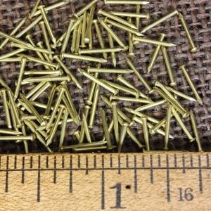 May include: A close-up of a pile of small, gold-colored tacks or brads. The tacks are scattered on a brown burlap surface. A ruler is placed below the tacks, showing a measurement of 1 inch.