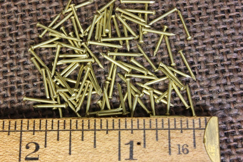 May include: A close-up of a pile of small, gold-colored tacks or brads. The tacks are scattered on a brown burlap surface. A ruler is placed below the tacks, showing a measurement of 1 inch.
