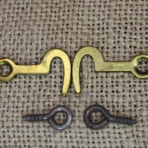 May include: A collection of hardware components, including two brass-colored hook-and-eye closures, two screws, and two eye screws. The hook-and-eye closures are approximately 1 inch long. The items are arranged on a textured, beige surface.