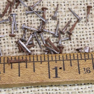 May include: A close-up of a wooden ruler with brass ends, marked with inches and fractions. The ruler is lying on a burlap surface with a scattering of small, rusty nails.