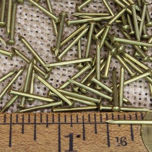 May include: A close-up of a pile of brass tacks, measuring 1 inch in length, on a burlap surface. A wooden ruler is placed below the tacks.