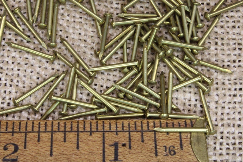 May include: A close-up of a pile of brass tacks, measuring 1 inch in length, on a burlap surface. A wooden ruler is placed below the tacks.