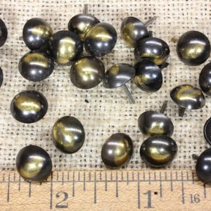 May include: A collection of 20 gold and black metal tacks, nail heads, or upholstery tacks. The tacks are arranged on a burlap surface with a ruler for scale.