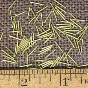 May include: A close-up of a pile of small, gold-colored tacks or nails on a brown burlap surface. A ruler is placed below the tacks, showing measurements in inches.
