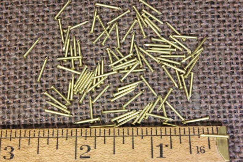 May include: A close-up of a pile of small, gold-colored tacks or nails on a brown burlap surface. A ruler is placed below the tacks, showing measurements in inches.