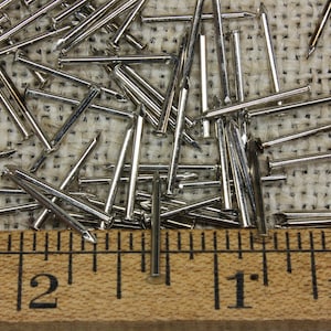 Könnte beinhalten: Nahaufnahme eines Stapels silberfarbener Reißnägel, die 2,5 cm lang sind, auf einer Jutestoffoberfläche. Ein Holzliniall mit Zollmarkierungen befindet sich am unteren Rand des Bildes.