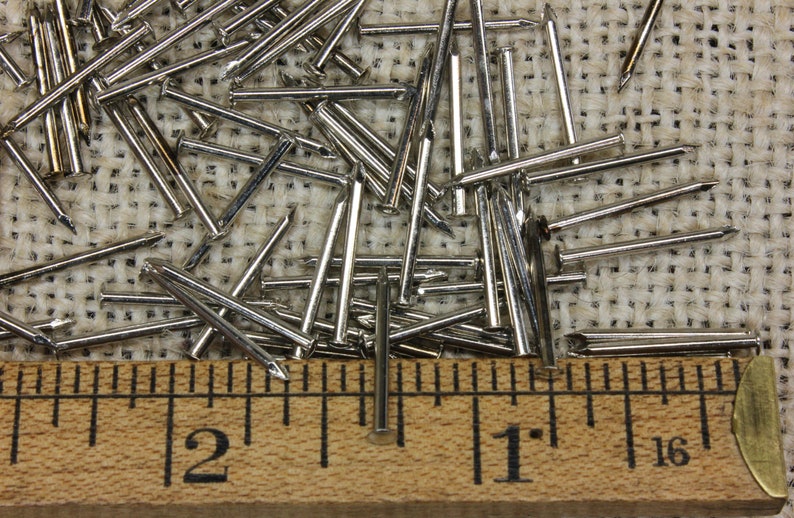 May include: A close-up of a pile of silver-colored tacks, measuring 1 inch in length, on a burlap surface. A wooden ruler with inch markings is positioned at the bottom of the image.